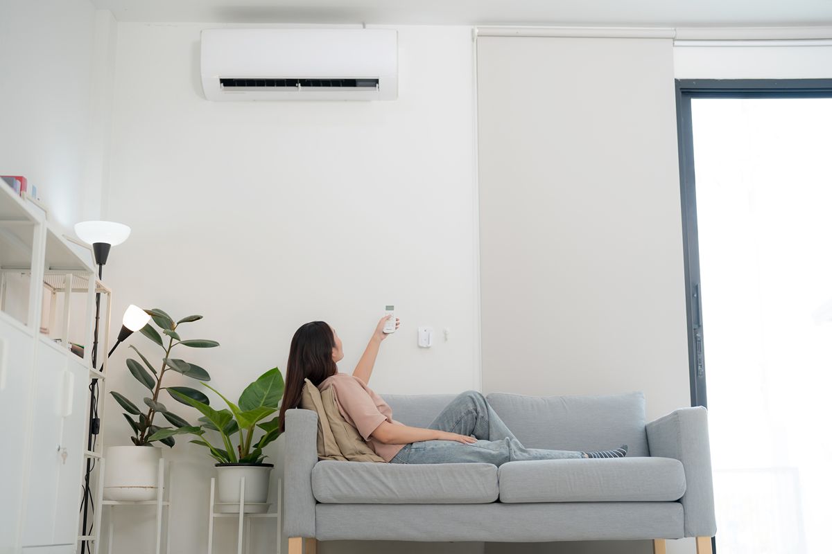 Woman relaxing on a sofa while using a remote control to adjust a wall-mounted split-type air conditioner