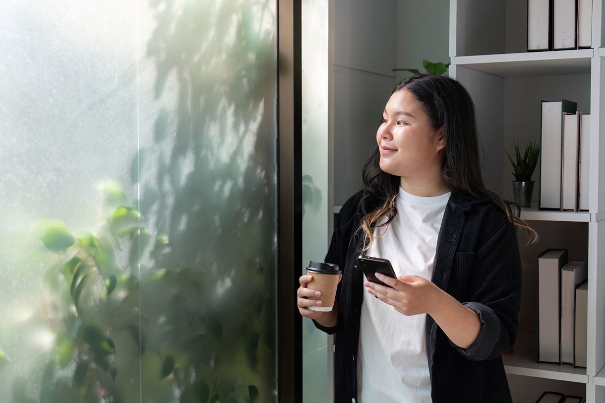 Woman holding a smartphone and coffee cup while looking through a rain-covered window at home