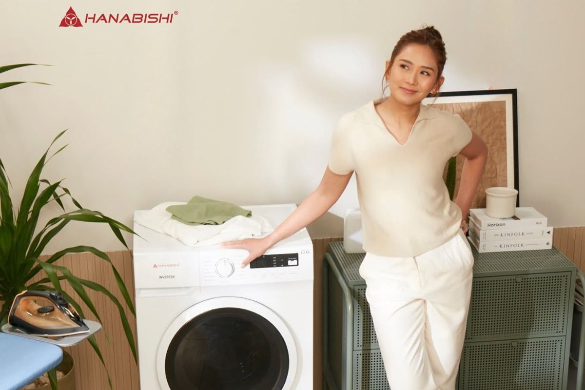 Sarah Geronimo standing beside a Hanabishi front-load washing machine in a home laundry area with an ironing board and storage cabinet